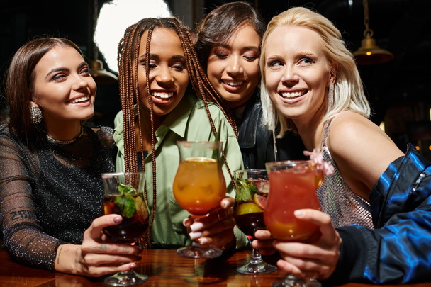 joyful and elegant female friends holding glasses with delicious cocktails in bar, friendship - Photo, Image
