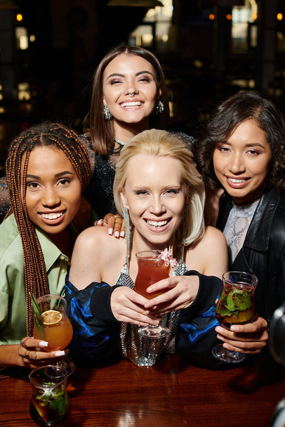 χαρούμενες και λαμπερές πολυπολιτισμικές γυναίκες με κοκτέιλ κοιτάζοντας κάμερα στο μπαρ, εορταστικό πάρτι - Φωτογραφία, εικόνα