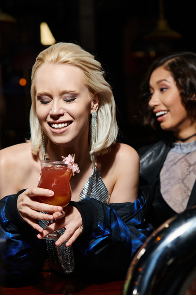 mujer rubia encantada con copa de delicioso cóctel cerca de novia multicultural en el bar - Foto, imagen