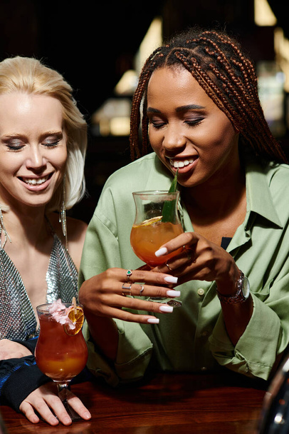 jovens e glamourosas namoradas multirraciais segurando óculos com coquetéis no bar, tempo de festa - Foto, Imagem