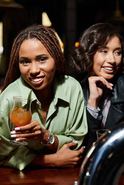 Barda Asyalı kız arkadaşının yanında kokteyl bardağıyla kameraya bakan şık Afro-Amerikan kadın. - Fotoğraf, Görsel