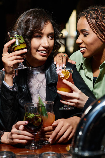 ammalianti ed eleganti fidanzate multirazziali sorridenti e con in mano deliziosi cocktail nel bar - Foto, immagini