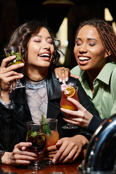 興奮するアフリカ系アメリカ人とアジアの女性 カクテルを保持し,バーで目を閉じて笑う - 写真・画像