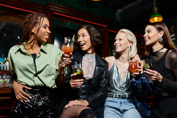 wesołe i stylowe wielonarodowe kobiety z pysznymi koktajlami rozmawiające w barze, wieczór panieński - Zdjęcie, obraz