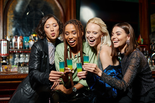 joyful and stylish multiracial girlfriends clinking shot glasses during festive party in bar - Photo, Image