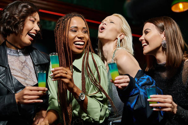 overjoyed and cheerful multicultural girlfriends holding shot glasses in bar, festive party - Photo, Image