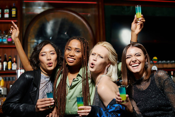 excited multiethnic girlfriends pouting lips and looking at camera in bar, glamorous lifestyle - Photo, Image