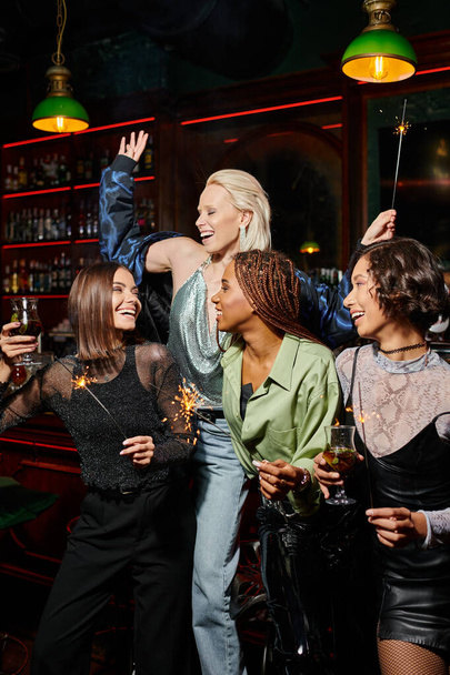 переполненные радостью многонациональные девушки с коктейлями и сверкающими искрами, веселящиеся в баре, время вечеринок - Фото, изображение