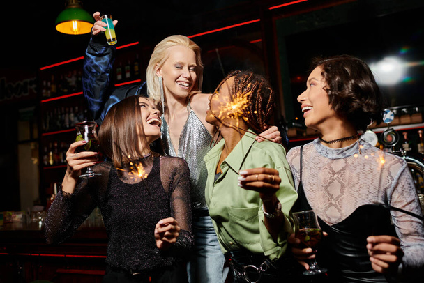 joyeuses et élégantes copines multiethniques avec des cocktails et des étincelles festives pendant la fête au bar - Photo, image