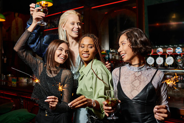 joyeuses petites amies multiethniques trinquant avec des cocktails et tenant des étincelles brillantes dans le bar, fête - Photo, image