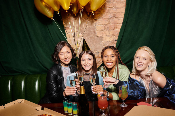 podekscytowane wielonarodowe dziewczyny wznoszące toast szampanem w pobliżu złotych balonów w barze, przyjęcie urodzinowe - Zdjęcie, obraz