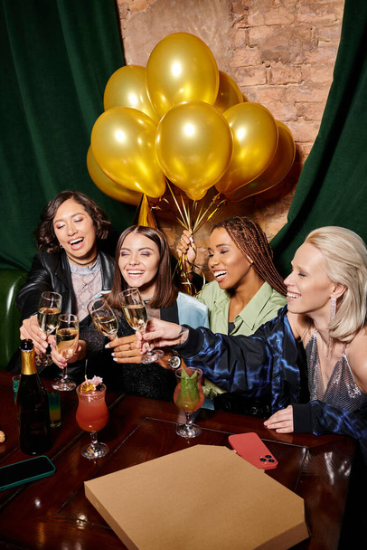 többnemzetiségű barátnők csattogó pezsgő poharak közelében arany lufik a bárban, születésnapi party - Fotó, kép