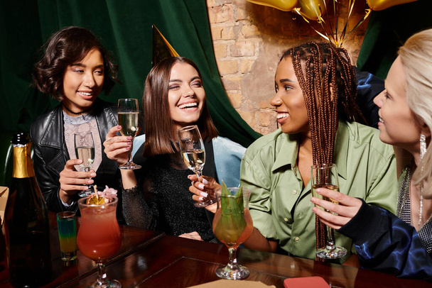 jong verjaardag meisje in party hoed houden champagne in de buurt multi-etnische vriendinnen en cocktails in bar - Foto, afbeelding