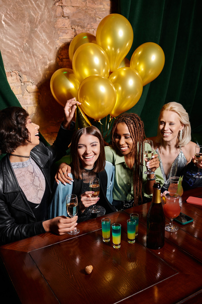 счастливые стильные мультиэтнические женщины с шампанским рядом с золотыми воздушными шарами и коктейлями в баре, день рождения - Фото, изображение