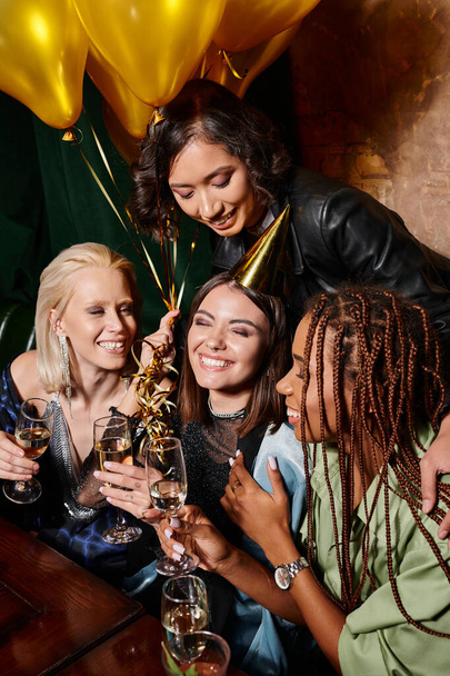 tevreden vrouw in feestmuts met champagne glimlachen met gesloten ogen in de buurt van multi-etnische vriendinnen - Foto, afbeelding