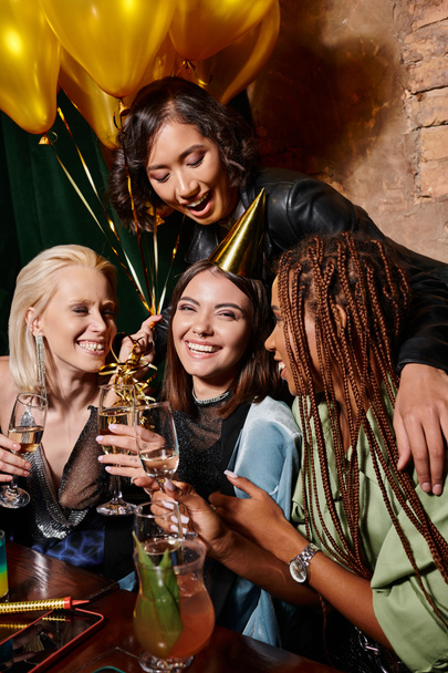 alegre mujer en partido sombrero celebración champán cerca multiétnica novias y globos en bar - Foto, Imagen
