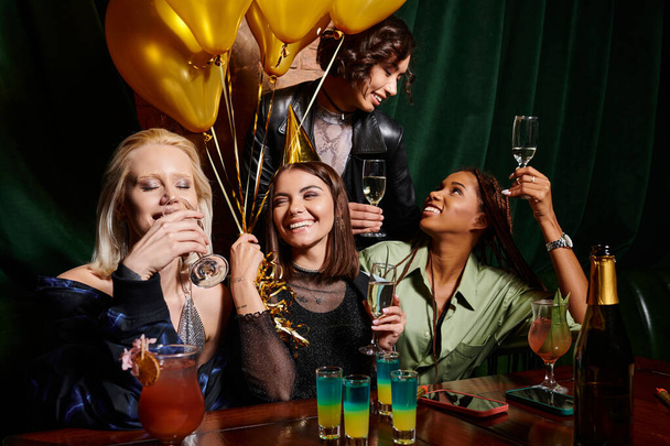 захоплені багаторасові подружки п'ють шампанське біля золотих кульок в барі, вечірка на день народження - Фото, зображення