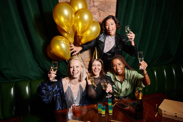 cheerful multicultural girlfriends with champagne looking at camera and celebrating birthday in bar - Photo, Image