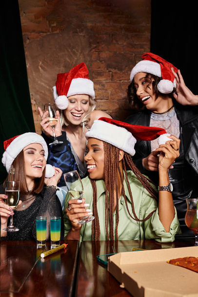 молодые и беззаботные многонациональные девушки в рождественских шляпах с шампанским, веселящиеся в баре, вечеринки - Фото, изображение