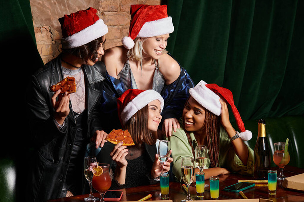 amigas multiétnicas felices en sombreros de Navidad celebrando el Año Nuevo con champán y pizza en el bar - Foto, Imagen