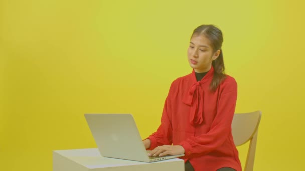 jonge vrouw het dragen van een rood shirt werken op laptop terwijl zitten op gele studio muur achtergrond. - Video