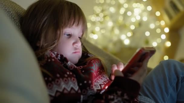 Uma criança assiste desenhos animados de Natal no telefone em casa. Uma rapariga está à espera do Pai Natal. Imagens 4k de alta qualidade - Filmagem, Vídeo