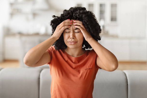 Headache Concept. Young black woman suffering from migraine at home, sick african american female rubbing temples while sitting on couch in living room, feeling despair, having acute pain, - Photo, Image