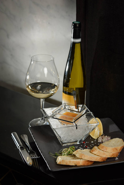 Pike hal kaviár, jégen, krutonnal és vajjal, átlátszó edényen, egy pohár fehér borral, sötét alapon - Fotó, kép