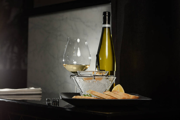 Caviar de poisson de brochet, sur glace, avec croûtons et beurre, sur un plat transparent avec un verre de vin blanc sur fond sombre - Photo, image