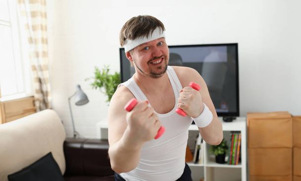 Молодой привлекательный мужчина занимается фитнесом дома держа гантель в руке, пытаясь бороться с лишним весом и потерей
 - Фото, изображение