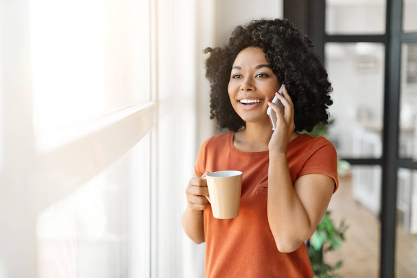 Belle jeune femme noire buvant du café et parlant sur un téléphone portable près de la fenêtre à la maison, souriant afro-américaine ayant agréable conversation mobile et profiter d'une boisson chaude, espace de copie - Photo, image