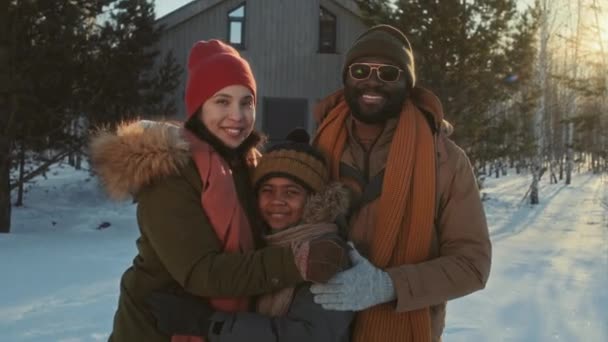 Середній портрет етнічно різноманітної пари та їх сина, що стоїть на відкритому повітрі на тлі сучасного дерев'яного будинку в зимовий день посміхаючись на камеру - Кадри, відео