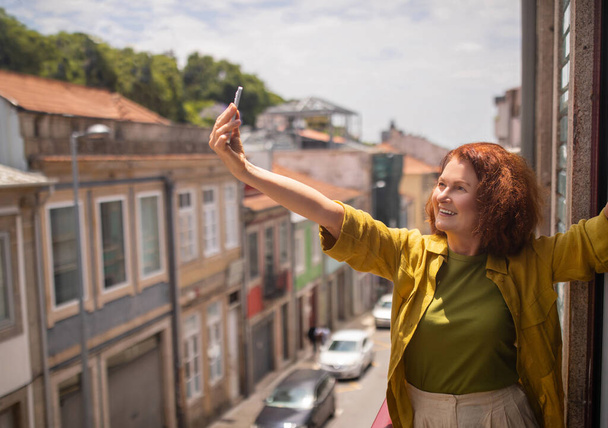 Joyful senior woman stretching out her arm and taking selfie on balcony with urban street view, smiling elderly female using smartphone, capturing photos at home, posing at cellphone camera - Photo, Image