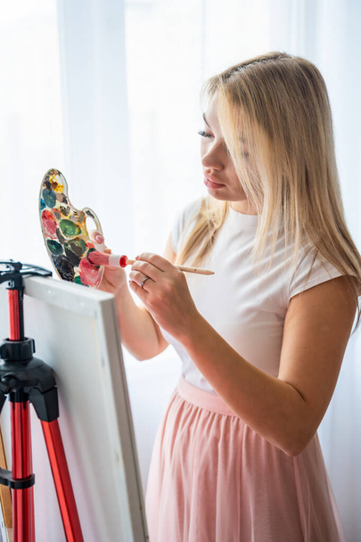 Paletli ve fırçalı güzel genç kadın ressam pencerenin yanındaki tuvale soyut pembe resim çiziyor. Sanat ve yaratıcılık kavramı. Yüksek kalite fotoğraf - Fotoğraf, Görsel