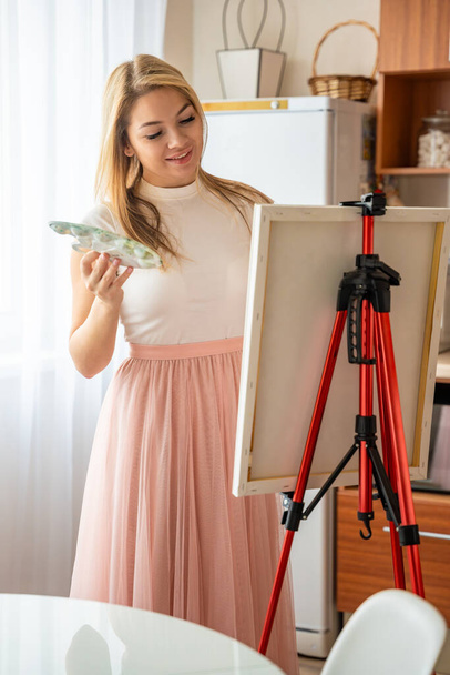 Krásná mladá blondýnka umělkyně s paletou a štětcem malování abstraktní růžový obrázek na plátně doma. Koncept umění a tvořivosti. Kvalitní fotografie - Fotografie, Obrázek