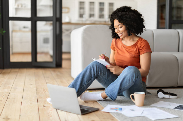 Mosolygó fiatal fekete nő diák segítségével laptop otthon és jegyzeteket, boldog afrikai amerikai nő tanulmány számítógép, miközben ül a padlón a nappaliban, élvezi az online oktatás, szabad tér - Fotó, kép