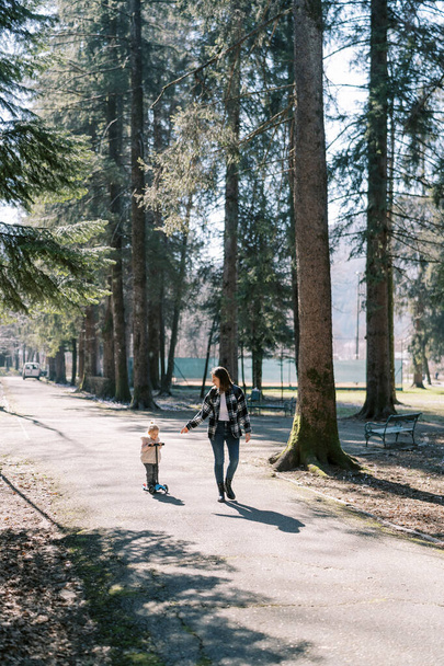 Anya egy kislány mellett sétál, aki robogózik az úton a parkban. Kiváló minőségű fénykép - Fotó, kép