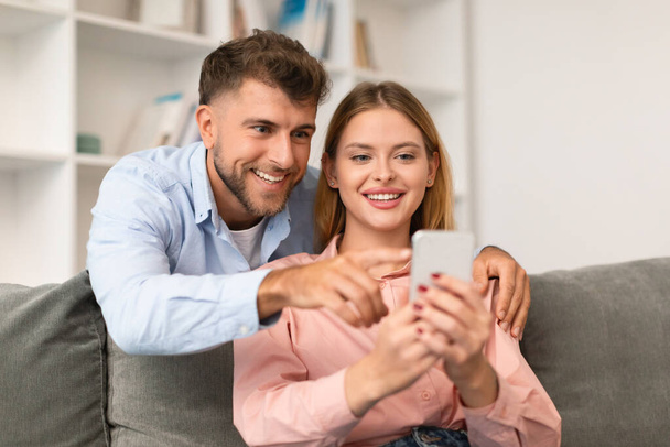 Mladí manželé používají mobilní telefon společně, procházejí aplikace a webové stránky, ukazují a sdílejí on-line nálezy, sedí spolu na gauči doma, zapojeni do digitálního zařízení - Fotografie, Obrázek
