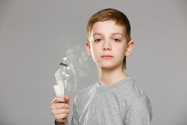 Unhappy boy holding inhalator mask releasing smoke on grey backg - Fotó, kép