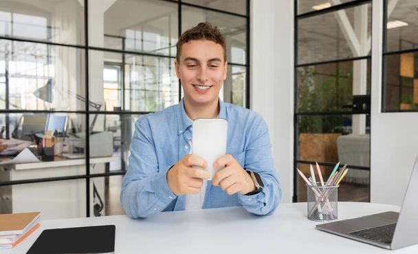 Усміхнений привабливий тисячолітній кавказький менеджер хлопець з ноутбуком за столом спілкування по смартфонах в сучасному коворкінгу. Робота з гаджетом, бізнес-додатком, професійна кар'єра - Фото, зображення