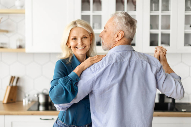Reife Romantik. Glückliches Seniorenpaar, das sich zu Hause umarmt und tanzt, Liebender älterer Mann und Frau, die zusammen in der Küche Spaß haben, Zeit miteinander genießen, Frau, die in die Kamera schaut, Nahaufnahme - Foto, Bild