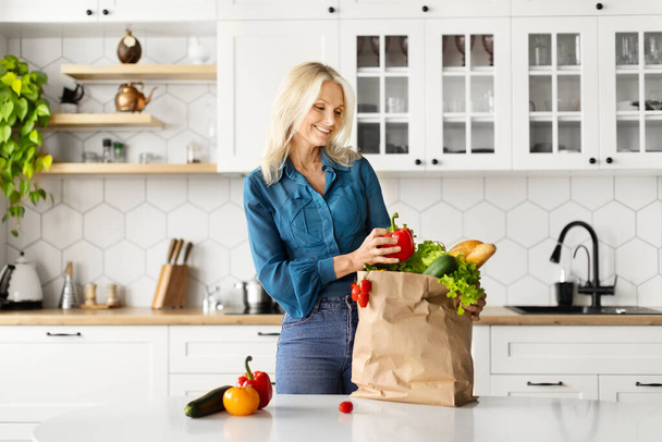Χαρούμενη ηλικιωμένη γυναίκα ξεπακετάρει χάρτινη σακούλα γεμάτη βιολογικά λαχανικά στην κουζίνα, χαρούμενη ηλικιωμένη κυρία που τσεκάρει φρέσκα λαχανικά μετά τα ψώνια φαγητού, στέκεται κοντά στο τραπέζι στο σπίτι, αντιγράφει χώρο - Φωτογραφία, εικόνα