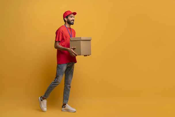 Livreur souriant en uniforme rouge et capuchon portant une boîte en carton, beau livreur heureux avec paquet dans les mains marchant sur fond jaune vif en studio, services de livraison de publicité - Photo, image