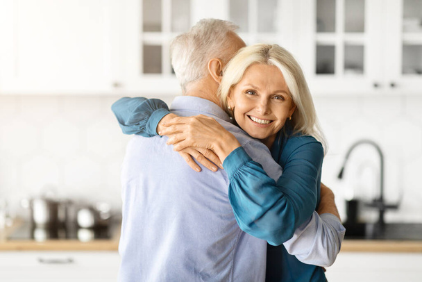Portret romantycznych starszych małżonków przytulających się we wnętrzu kuchni, kochający starszego mężczyznę i kobietę bawiących się razem w domu, nawiązywanie więzi i uśmiech, piękna starsza kobieta patrząca przed kamerą, wolna przestrzeń - Zdjęcie, obraz