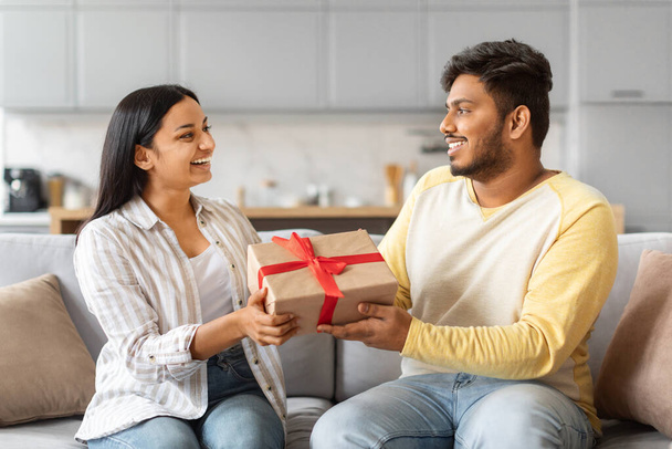 Happy young Indian couple exchanging gifts while sitting on sofa at home, loving man giving beautifully wrapped present with red ribbon to his excited girlfriend, joyful spouses greeting each other - Photo, Image