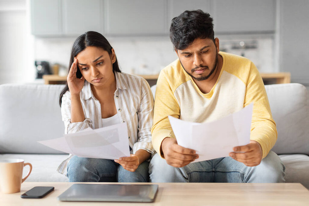 Stresszes fiatal indiai házastársak, akik otthon ellenőrzik a pénzügyi papírokat, feldúlt keleti házaspár ül a kanapén és hiteldokumentumokat olvas, számlákat tart, havi kiadásokat számol, családi költségvetést tervez - Fotó, kép