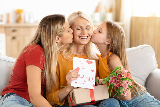 Liebevolle erwachsene Tochter und frühzeitige Enkelin küssen und umarmen, Oma mit Muttertag grüßen, aufgeregte ältere Dame hält Geschenk, Blumen, handgemachte Grußkarte - Foto, Bild
