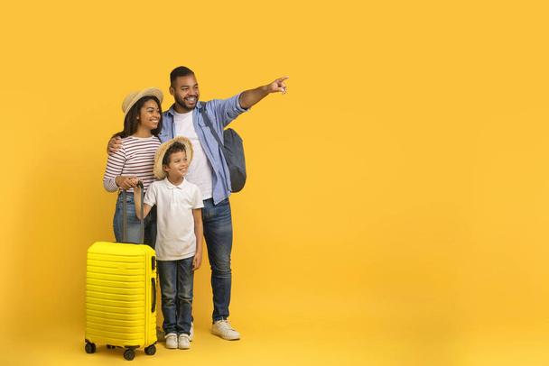 Familienreisekonzept. Glückliche afrikanisch-amerikanische Eltern mit kleinem Sohn, der Koffer trägt und auf Kopierraum auf gelbem Hintergrund zeigt, Mama, Papa und männliches Kind urlaubsreif, Panorama - Foto, Bild