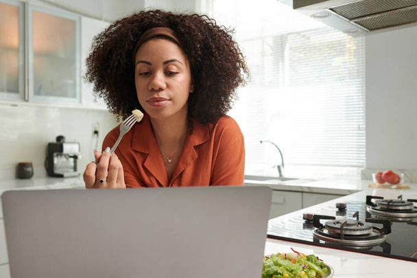 İş kadını mutfak tezgahında salata yiyor ve e-posta ya da dizüstü bilgisayardaki makaleleri okuyor. - Fotoğraf, Görsel