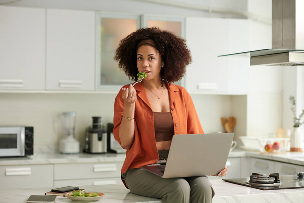 Portret van een vrouw op het aanrecht, werkend op een laptop en salade etend - Foto, afbeelding
